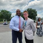 Two people shaking hands outdoors with the U.S. Capitol in the background. A person stands nearby. Trees and clouds complete the scene.