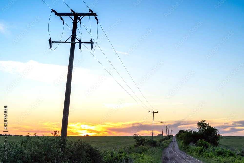 A dirt road runs alongside power lines at sunrise or sunset in a rural landscape, with open fields and scattered bushes.