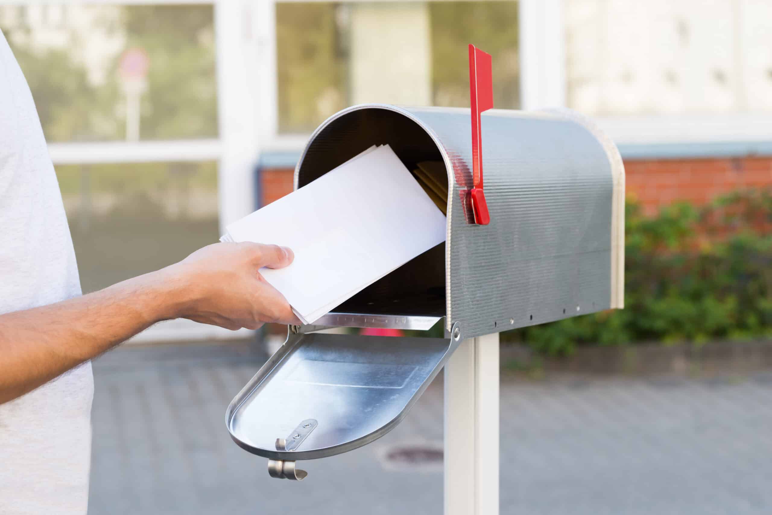 A person places envelopes into a silver mailbox with a red flag, situated outdoors in front of a building.