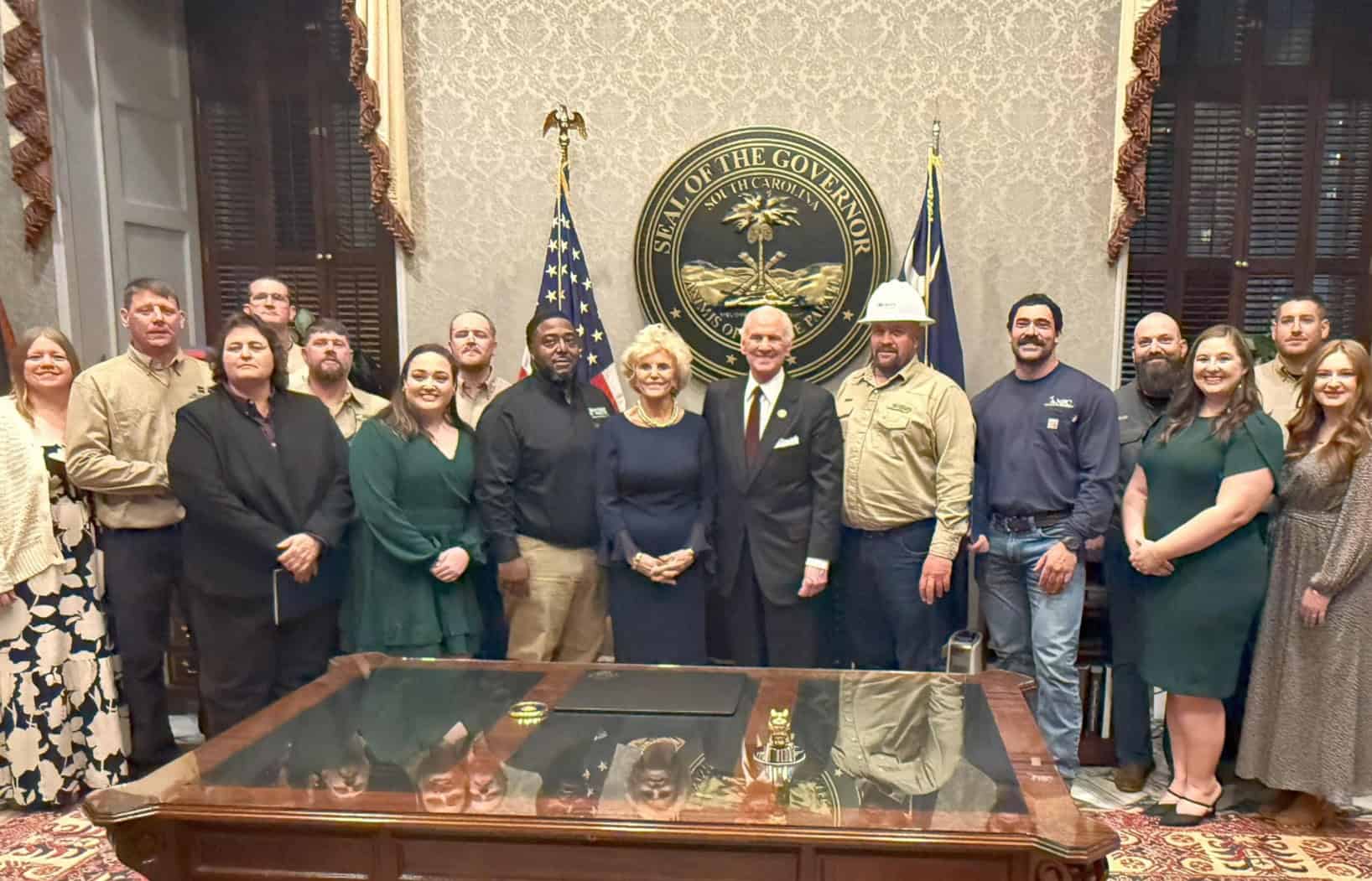 A group of people stands in front of the South Carolina Governor's seal, within an ornate room featuring flags and a wooden table.