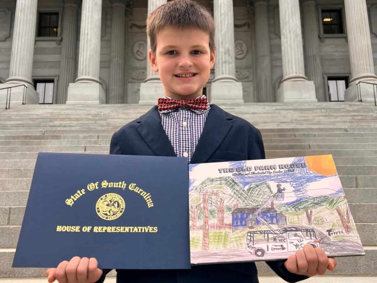 A person holds a certificate and drawing outside the South Carolina State House, standing on steps with large columns in the background.