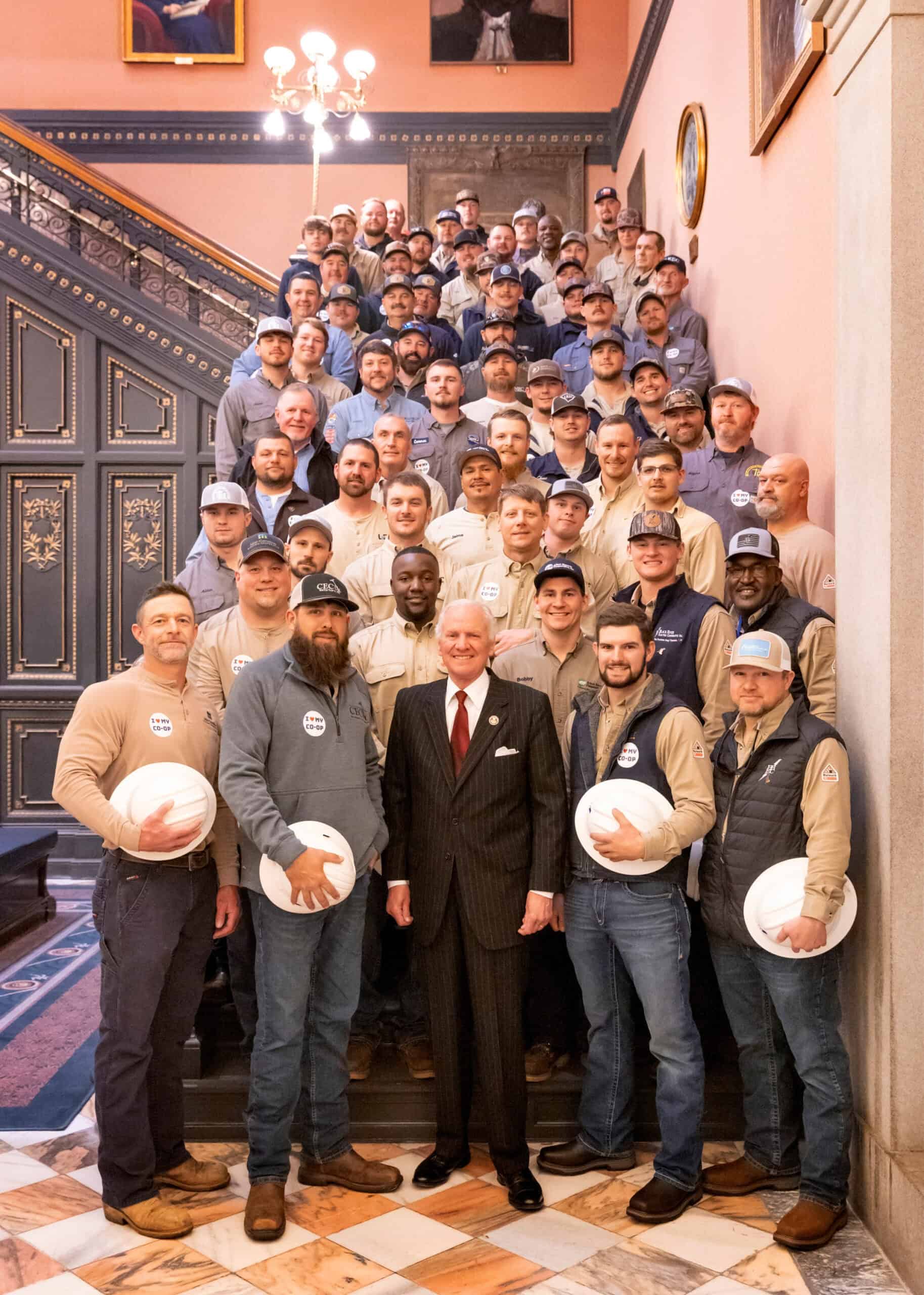 A group of people, some holding white hard hats, stands on a staircase in a historic building with ornate decorations and framed portraits.