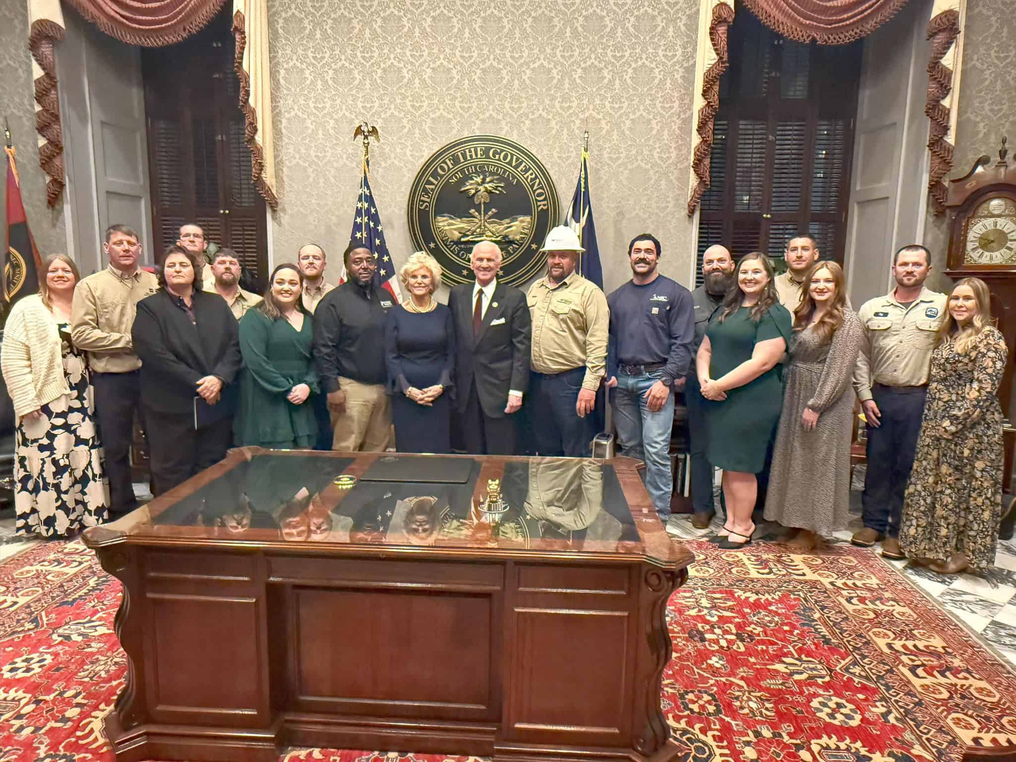 A group of people stands in an ornate room with a desk, featuring the Seal of the Governor of South Carolina.