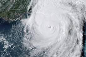 Aerial view of a massive hurricane swirling over water, with visible eye and cloud bands approaching land, indicating potential impact on coastal areas.