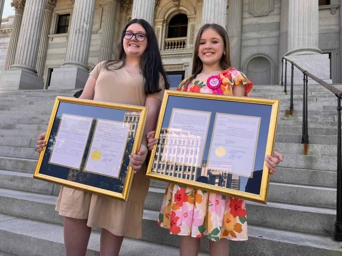 Two children hold framed certificates on stone steps, outside a historic building with tall columns. They appear happy and proud.