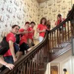 A group of people in red shirts pose on a wooden staircase with ornate wallpapered walls in a historic setting.