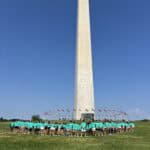 People in matching teal shirts stand in front of the Washington Monument, surrounded by flags under a clear blue sky.