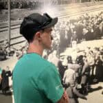 A person in a green shirt and cap observes a historical black-and-white photograph depicting a large crowd near train tracks.