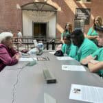 A person speaks to a group of people at a table in the United States Holocaust Memorial Museum's exhibit area. People listen attentively.