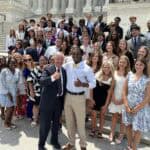 A group of people posing on the steps of the U.S. Capitol building, smiling and dressed in formal attire, with one person giving thumbs up.