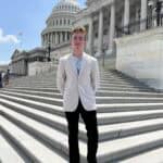 A person stands on the steps of the U.S. Capitol, with the iconic dome visible under a clear blue sky.