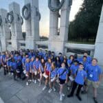 A group of people in blue shirts pose at the World War II Memorial in Washington, D.C., surrounded by state pillar monuments.