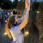 A person traces names on the Vietnam Veterans Memorial wall at dusk, surrounded by people, with trees and lights in the background.