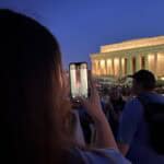 A person photographs the illuminated Lincoln Memorial at night, capturing architectural details. Crowds gather around, enjoying the iconic landmark in Washington, D.C.