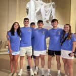 Six people in matching blue shirts pose at the Lincoln Memorial, featuring the prominent statue of Abraham Lincoln.