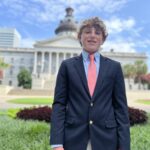 A young person in a suit stands in front of the South Carolina State House, with a clear blue sky background.