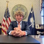 A person seated at a desk in an official office with American and state flags, seal on wall, framed pictures behind.