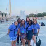 Six people in matching blue shirts pose near the World War II Memorial fountain, with the Washington Monument visible in the background.