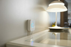 A smart home device is mounted on a wall in a minimalistic room, next to a modern table lamp on a glass surface.