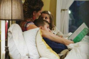 A person reads a book to two children on a bed, under warm lighting, with a lamp and window in the background.