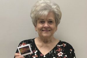 A person with short white hair smiles while holding a "congrats" card, wearing a black floral top against a plain background.