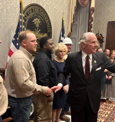 Several people gather in a formal room with flags. The South Carolina Governor's seal is visible on the wall in the background.