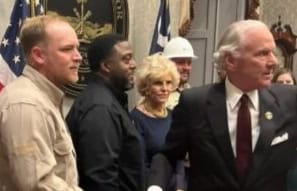 A group of people, including one in a hard hat, gathered indoors at the South Carolina Statehouse with the Governor near flags and a wall plaque with an emblem design.