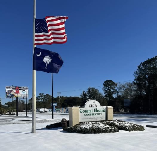 Two flags fly above a snow-covered sign reading "Coastal Electric Cooperative." Trees and a billboard are visible in the background.