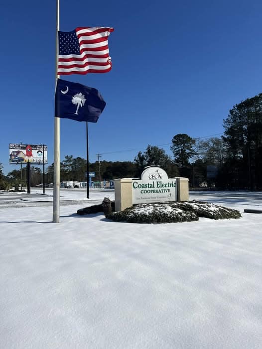 Two flags fly above a snow-covered sign reading "Coastal Electric Cooperative." Trees and a billboard are visible in the background.